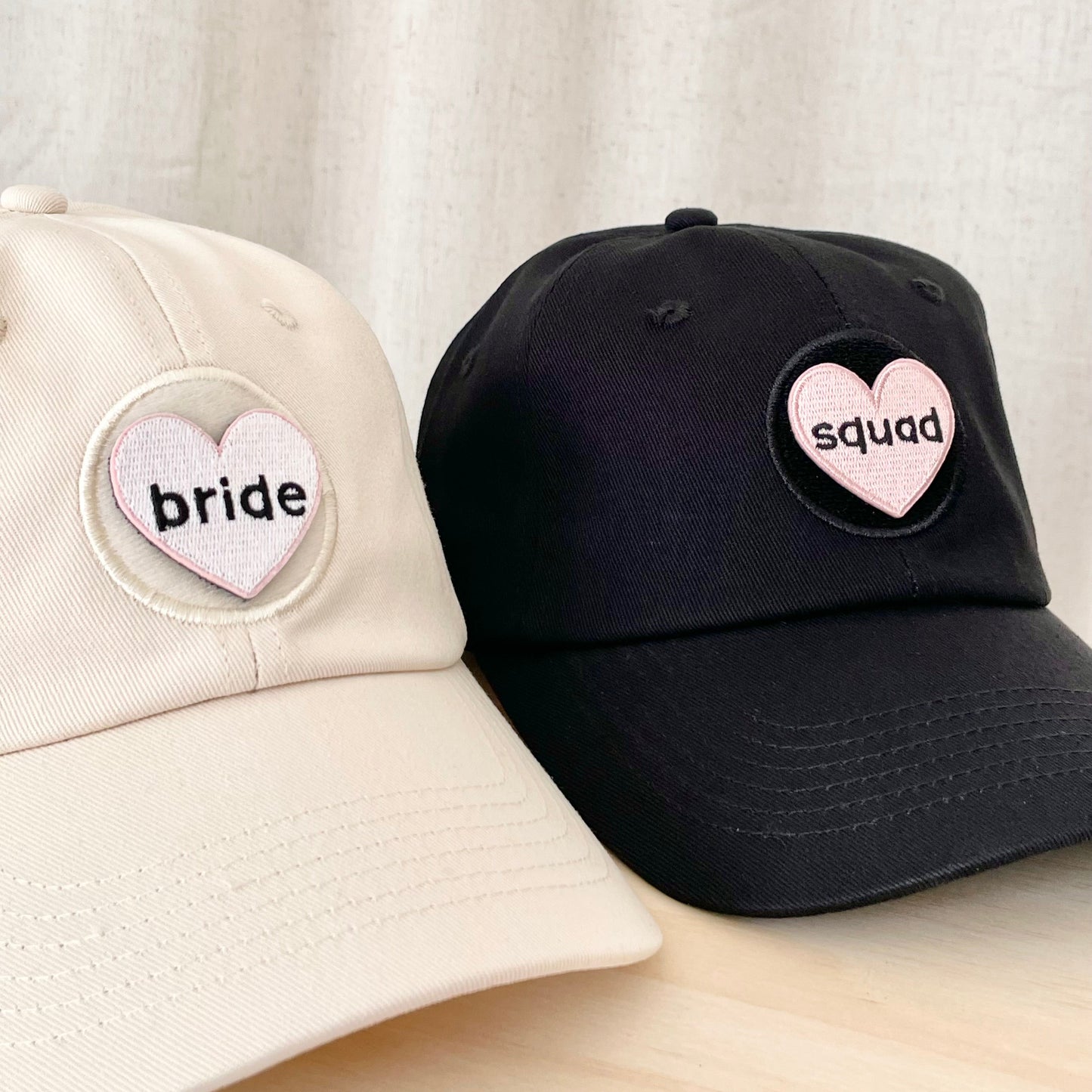 a cream hat with a white heart shaped velcro patch with black text that says "bride" next to a black hat with a pink heart shaped velcro patch with black text that says "squad"