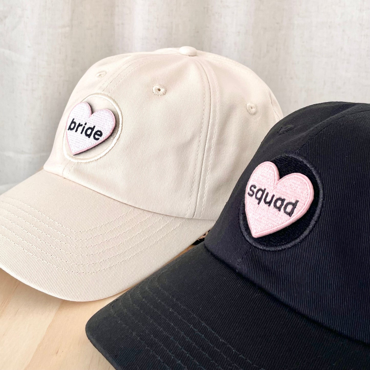 side view of a cream hat with a heart shaped velcro patch that says "bride" next to a black hat with heart shaped velcro patch that says "squad"