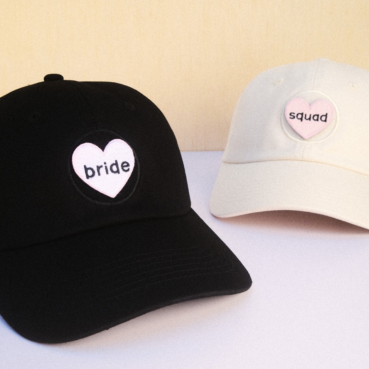 Black cap with 'bride' heart design and beige cap with 'squad' heart design on a light background