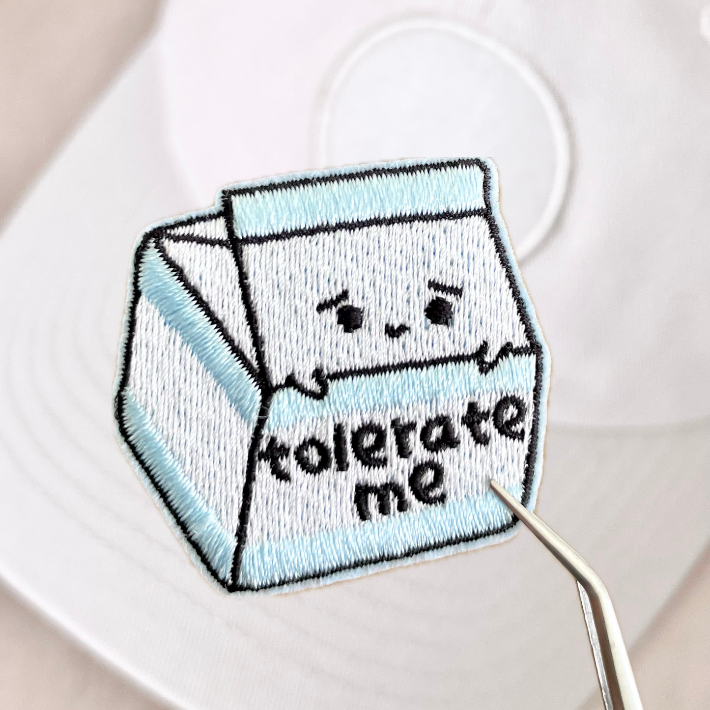 A velcro patch designed to look like a lactose-free milk carton with the phrase 'tolerate me' on it, held against a white background.