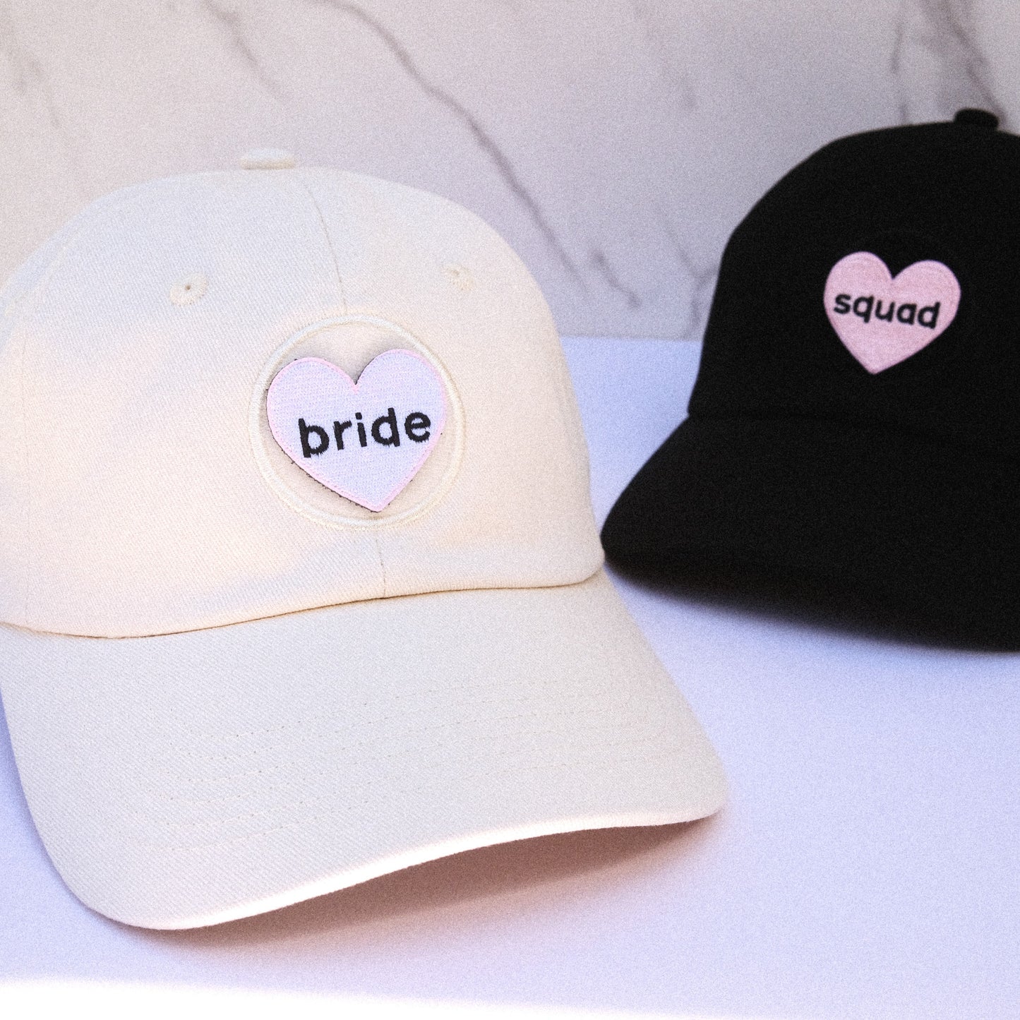 Two baseball caps, one white and one black, with heart-shaped velcro patches that say "bride" and "squad" respectively on a light background.